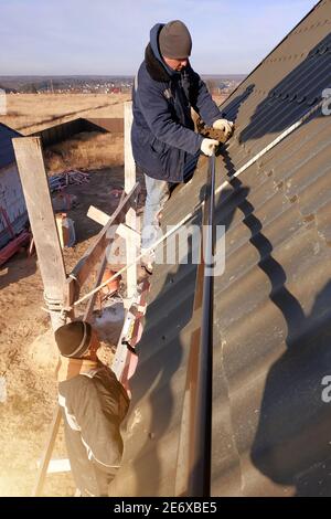 Il padrone lavora sul tetto di una casa privata senza assicurazione, il lavoro è eseguito nella stagione fredda, il lavoratore si comporta con attenzione, il tetto è Foto Stock