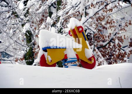 Moto dondolante in primavera, giocattolo in legno rosso e giallo in giardino in inverno, con alberi innevati sullo sfondo in estrema nevicata. Foto Stock