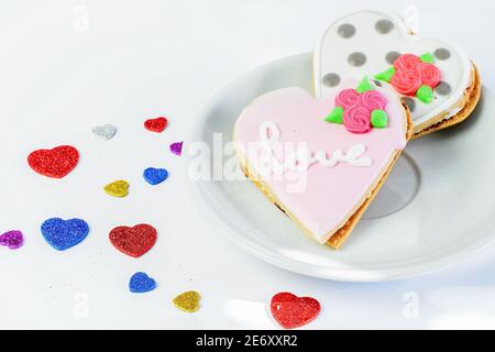 Due dolci torte glassate in un da di cuori sul piatto bianco e sullo sfondo bianco con alcuni piccoli cuori colorati primo piano. Simbolo di due amore Foto Stock