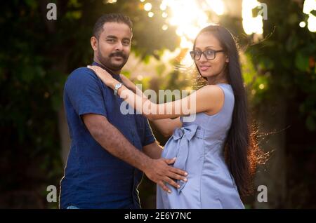 Giovane coppia che si tiene l'un l'altro in serata all'aperto, il sole che tramonta attraverso gli alberi sullo sfondo, una coppia incinta in attesa di un bambino presto, puzzare Foto Stock