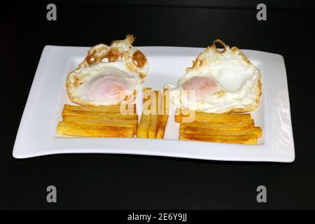 Due uova fritte con patatine fritte tagliate a bastoncini regolari e poste su un piatto rettangolare con fondo nero. Foto Stock