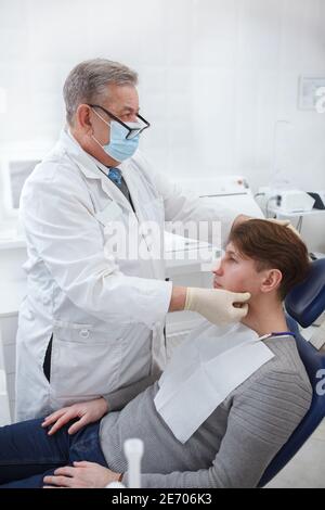 Immagine verticale di un dentista professionista che indossa una maschera medica per il viso, esaminando i nods linfatici del paziente prima del trattamento dentale Foto Stock
