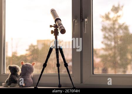 sulla soglia della finestra si trova su un treppiede un telescopio che punta al cielo, seduto accanto a due giocattoli morbidi girati per la strada, marrone tinta, retro, auto-i Foto Stock
