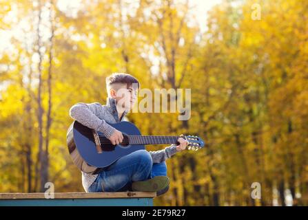 teen boy suona la chitarra acustica nera in un giorno d'autunno Nel Parco Foto Stock