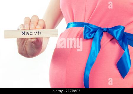 Donna incinta in abito rosa con nastro blu che mostra la parola marcia scritta su cubo di legno. Attesa per neonato ed espansione della famiglia Foto Stock