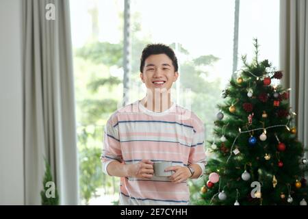 fuoco selettivo dell'uomo allegro che tiene la tazza vicino all'albero di natale in soggiorno Foto Stock