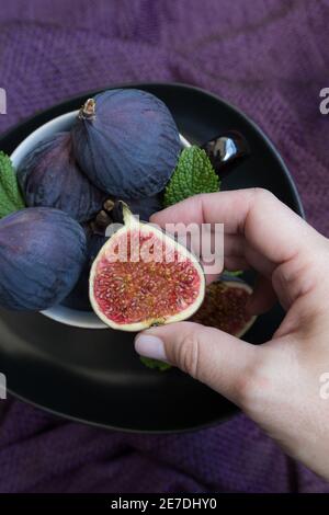 Vista in primo piano di una mano di donna che prende un fico dimezzato pieno di semi colorati. Altri tre fichi interi sono all'interno di una tazza. Foto Stock