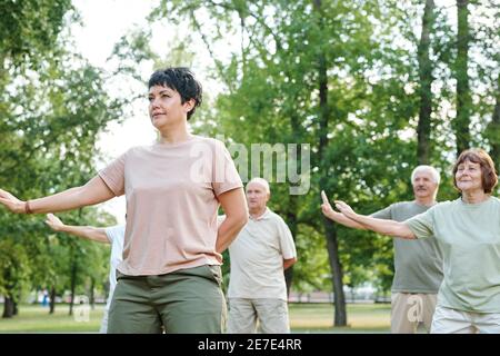 Donna matura allenarsi all'aperto nel parco con altri maturi persone sullo sfondo Foto Stock