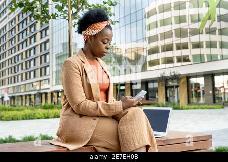 Donna d'affari africana seduta sulla panchina e digitando un messaggio il suo telefono cellulare all'aperto in città Foto Stock