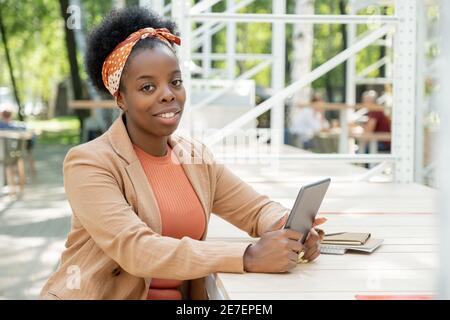 Ritratto di donna africana che sorride alla macchina fotografica mentre si siede a. il tavolo in caffetteria e utilizzando un tablet digitale Foto Stock