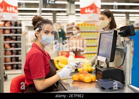 Giovane donna cassiere in guanti protettivi, maschera e schermo scansione confezione di arance fresche sopra il bancone mentre servire uno dei clienti Foto Stock