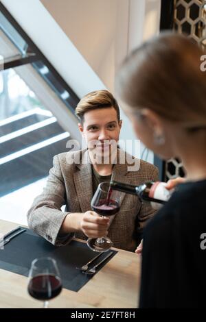 Elegante giovane seduto accanto al tavolo servito in un lussuoso ristorante e tenendo in mano l'eneglass mentre la cameriera versa in essa il vino rosso Foto Stock