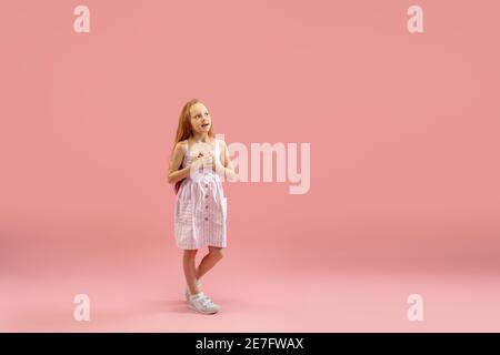 Sognando, si chiedeva. Infanzia e sogno sul grande e famoso futuro. Bella ragazza longhair su sfondo corallo rosa studio. Infanzia, sogni, immaginazione, educazione, espressione facciale, concetto di emozioni. Foto Stock