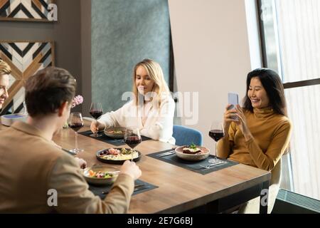Giovane donna asiatica felice con smartphone che scatta foto di ragazzi a cena mentre si siede al tavolo servito nel ristorante tra i suoi amici Foto Stock