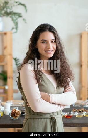 Giovane donna bruna sorridente che attraversa le braccia dal petto mentre si è in piedi contro tavola con ingredienti per la produzione di prodotti cosmetici naturali Foto Stock