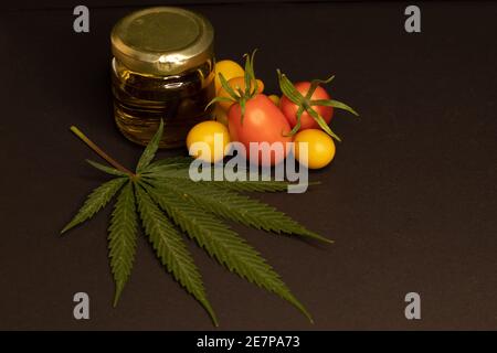 Una foglia di cannabis, un vaso pieno di olio cbd e alcuni pomodori ciliegini su sfondo nero. L'immagine raffigura il rapporto tra cibo e cannabis. Foto Stock