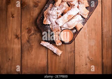 Cibo di Carnevale caffè TIPICO e aromatico su uno sfondo boscoso vista dall'alto. Sfrappole o chiachiere o ali d'angelo. Pasta dolce tradizionale fritta e cosparsa di zucchero in polvere. Foto Stock