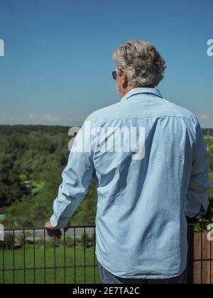 L'uomo anziano con i capelli grigi rimane in giardino e guarda all'orizzonte. Da dietro. Foto Stock