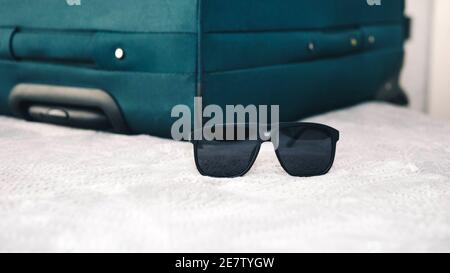 Oggetti da viaggio, occhiali da sole con valigia sul letto, concetto di viaggio, occhiali da sole neri dal sole. Foto Stock