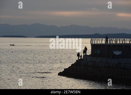 I pedoni dalle silhouette camminano lungo il frangiflutti di Ogden Point e guardano una piccola barca a motore al tramonto a Victoria sull'isola di Vancouver, British col Foto Stock