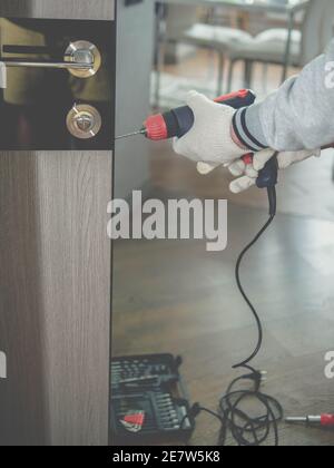 Un falegname ripara una porta, un installatore installa una porta interna in un appartamento, un giovane uomo trapina in una serratura della porta. Trapano elettrico nel processo. Foto Stock