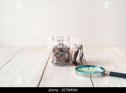 Soldi in un vaso di vetro e una lente d'ingrandimento accanto al vaso su uno sfondo chiaro. Foto Stock