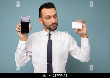 Bell'uomo che indossa abiti quotidiani isolati sulla parete di fondo di tenuta e usando il telefono e la carta di credito che fa il pagamento guardando carta bancaria in plastica Foto Stock