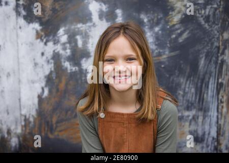 Giovane ragazza in un vestito marrone generale in inverno seduto vicino a un muro di un magazzino urbano Foto Stock