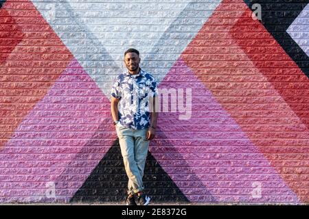 Un uomo nero si appoggia contro un colorato murale di strada e sorride alla telecamera. Foto Stock