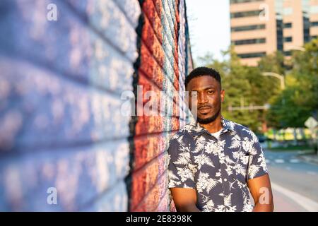 Un uomo nero si appoggia contro un colorato murale di strada e sorride alla telecamera. Foto Stock