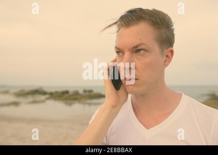 Chiusura del giovane uomo bello pensare mentre parlano al telefono cellulare presso la spiaggia pubblica di Hua Hin in Thailandia Foto Stock
