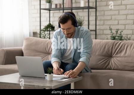 Uomo serio che indossa le cuffie scrivere note, studiando online a casa Foto Stock