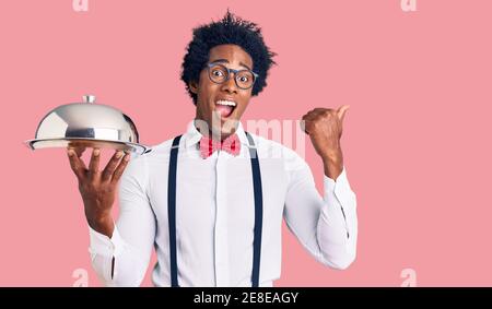 Bell'uomo afroamericano con capelli afro che indossa uniforme cameriera tenendo il vassoio argentato che punta il pollice verso il lato sorridente felice con bocca aperta Foto Stock