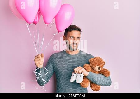 Bell'uomo con barba che si aspetta una bambina che tiene palloncini, scarpe e orsacchiotto sorridendo guardando al lato e fissando via pensando. Foto Stock