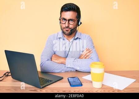 Giovane ispanico seduto alla scrivania indossando le cuffie dell'operatore presso l'ufficio del call center, scettico e nervoso, disapprovando l'espressione sul viso con cr Foto Stock