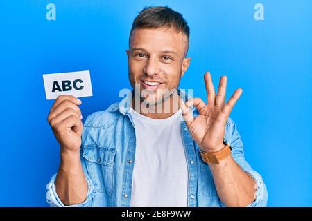 Bell'uomo del muscolo che tiene le lettere abc sulla carta che fa il segno ok con le dita, sorridente amichevole gesturing simbolo eccellente Foto Stock