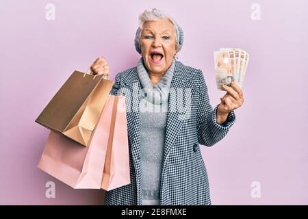 Donna anziana dai capelli grigi che tiene i sacchetti della spesa e le banconote delle libbre del regno unito che sorridono e ridono duro fuori forte perché divertente scherzo pazzo. Foto Stock