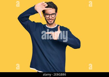 Giovane uomo ispanico che indossa abiti e occhiali casual sorridente rendendo cornice con mani e dita con volto felice. Creatività e concetto di fotografia. Foto Stock