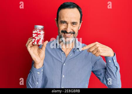 Uomo ispanico di mezza età che tiene il vaso di pillole che punta il dito ad uno sorridente felice e orgoglioso Foto Stock