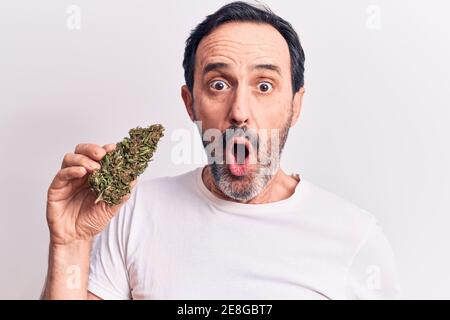 Bell'uomo di mezza età che tiene l'erbaccia di cannabis in piedi su uno sfondo bianco isolato spaventato e stupito con bocca aperta per sorpresa, viso incredulo Foto Stock