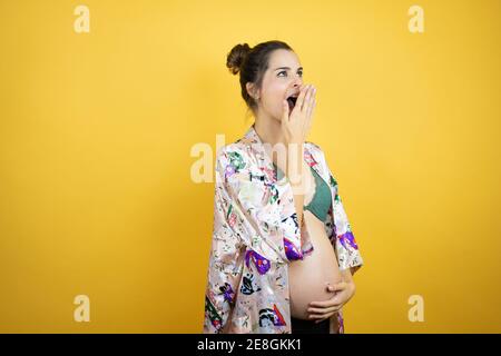 Giovane bella donna incinta aspettandosi bambino che indossa pajama su isolato sfondo giallo che copre la bocca con la mano, scioccato e impaurito per errore. Foto Stock