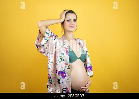 Giovane bella donna incinta aspettandosi bambino che indossa pajama su isolato sfondo giallo mettendo una mano sulla testa sorridendo come aveva dimenticato Foto Stock