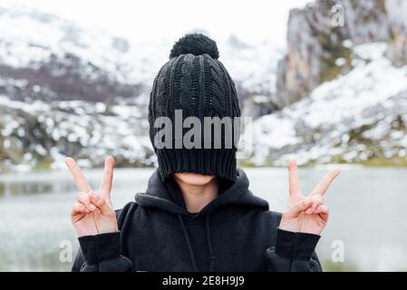 Anonimo femmina nascondendo faccia dietro cappello nero e mostrando v segno mentre si sta in piedi su altopiani innevati e accidentati sulla riva del lago Foto Stock