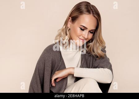 giovane donna caucasica indossare poncho guardando giù con sorriso mentre seduta sul pavimento in studio Foto Stock