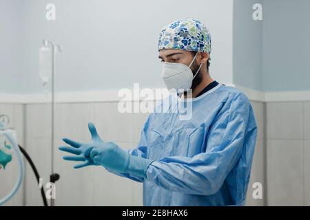 Anonimo giovane veterinario maschile in uniforme sterile mettendo su elastico guanti durante la preparazione all'intervento chirurgico in sala operatoria Foto Stock