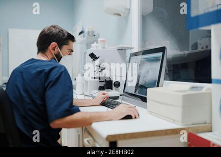 Vista posteriore del veterinario maschile focalizzato irriconoscibile in uniforme di osservazione Illustrazione a raggi X su computer desktop in laboratorio Foto Stock