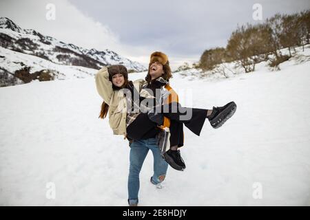 Giovane ridente viaggiatore maschio con occhi chiusi portando femmina amata Sul monte innevato sotto il cielo nuvoloso in Spagna Foto Stock