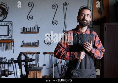 Contenuto giovane artigiano etnico in camicia a scacchi e grembiule tenendo il martello in mano e guardando la fotocamera durante il lavoro in miti Foto Stock