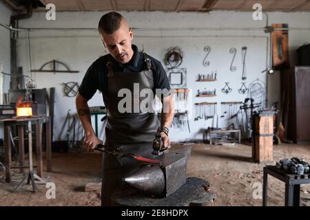 Vista frontale della giovane forger maschile tatuata in grembiule riscaldare il metallo in forno mentre si lavora in macerato smithy Foto Stock
