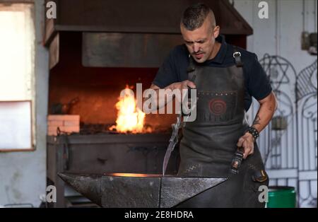 Concentrato giovane fabbro bearded in grembiule e occhiali di protezione colpisce metallo riscaldato con martello sull'incudine mentre si lavora in fucina Foto Stock